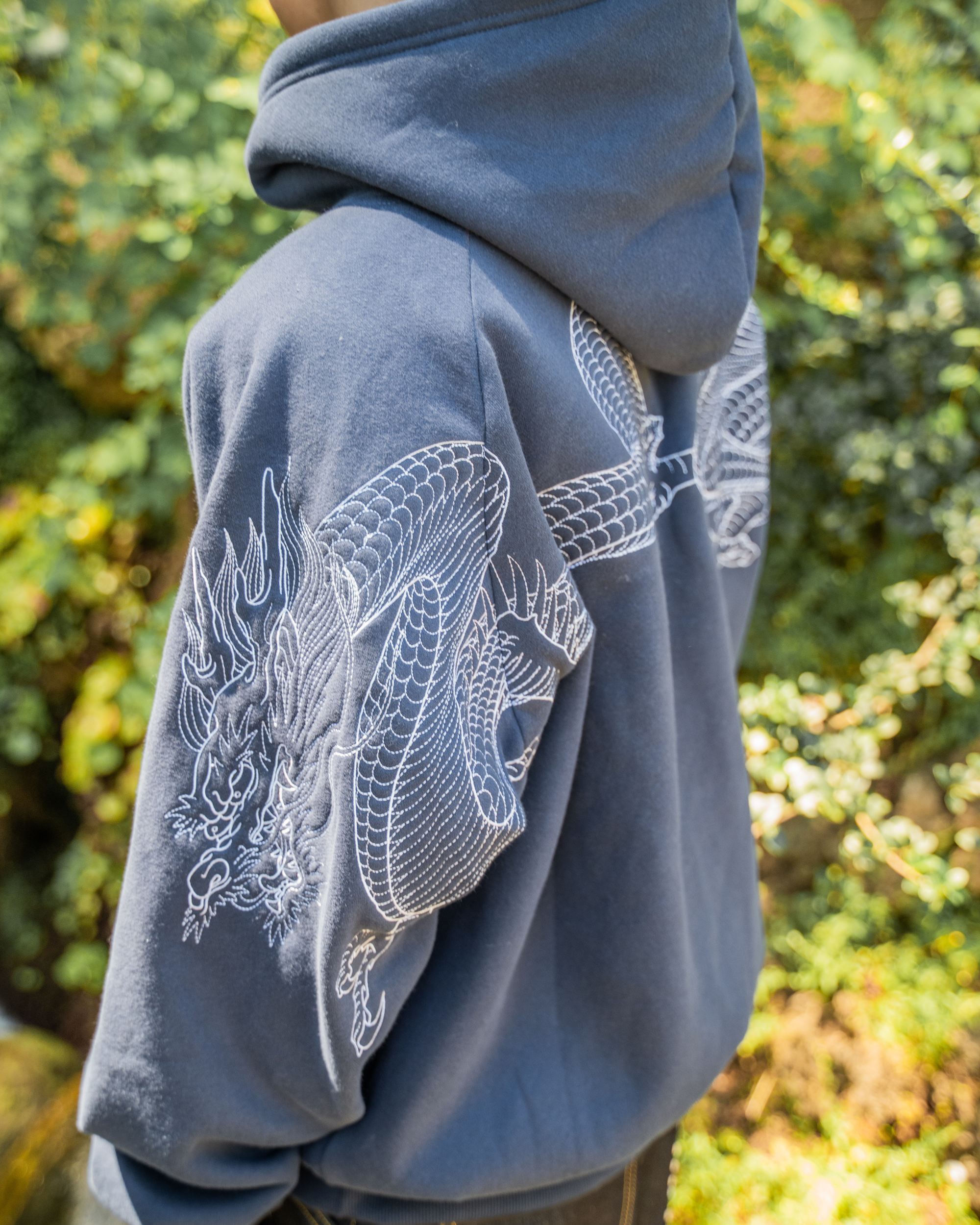 A person outdoors wearing a slate blue hoodie with large white embroidered dragon designs across the back and sleeves, set against a blurred green foliage background.