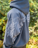 A person outdoors wearing a slate blue hoodie with large white embroidered dragon designs across the back and sleeves, set against a blurred green foliage background.