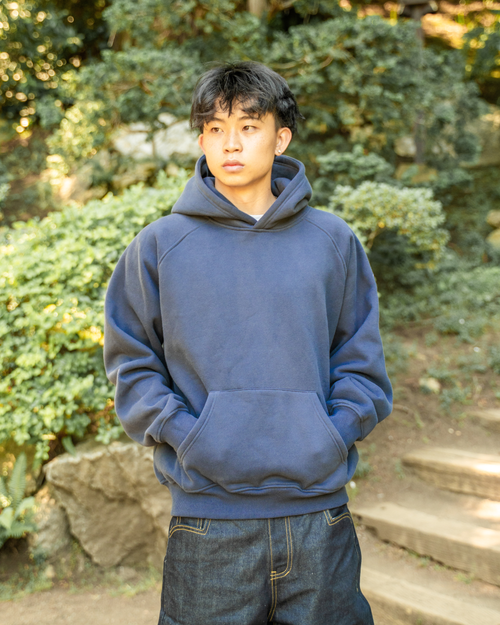 A young man wearing a slate blue pullover hoodie and dark blue jeans, standing outdoors in a garden setting with stone steps and green foliage in the background.