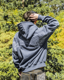 A person viewed from behind wearing a dark slate blue hoodie with a large white dragon graphic across the back, standing outdoors in front of a green forest background.